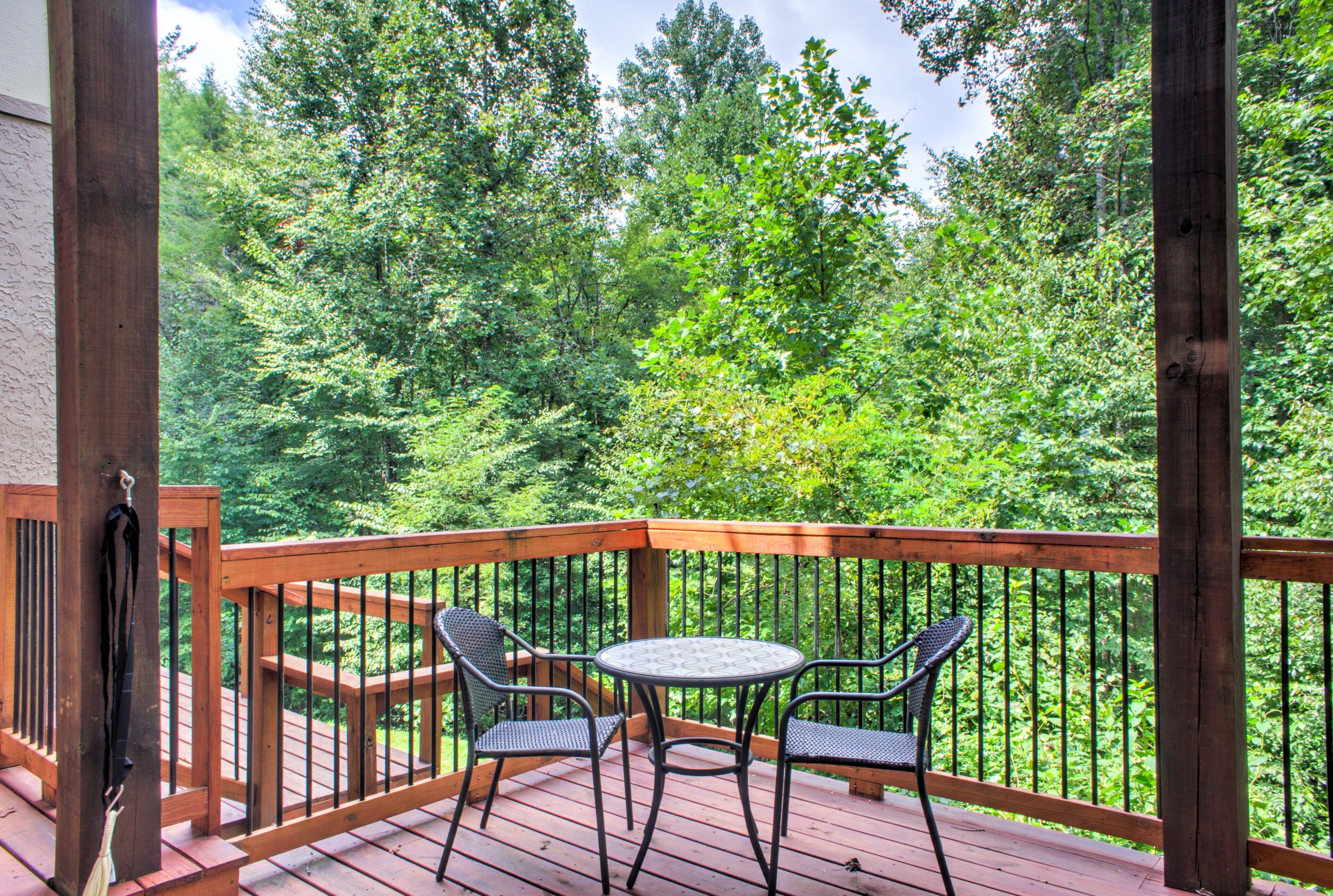 Una terraza de madera con una mesa redonda y dos sillas, con vista a un bosque frondoso.