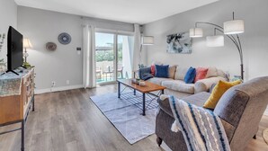 Un salon moderne avec un canapé, une table basse et un téléviseur posé sur un meuble en bois. On y trouve une grande fenêtre avec des rideaux, un lampadaire et des œuvres d’art accrochées au mur.