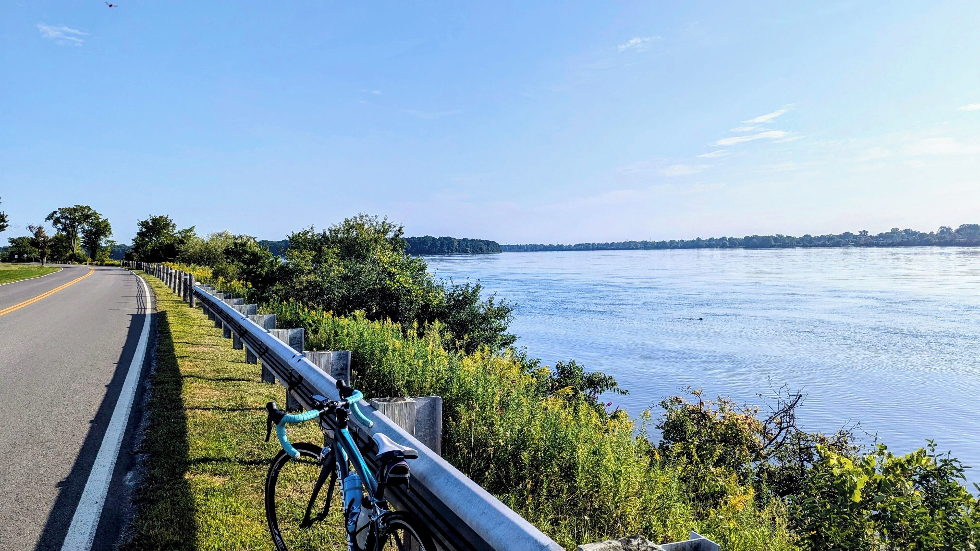 A road with a bicycle parked by the side, overlooking a calm body of water with trees and a distant shoreline.
