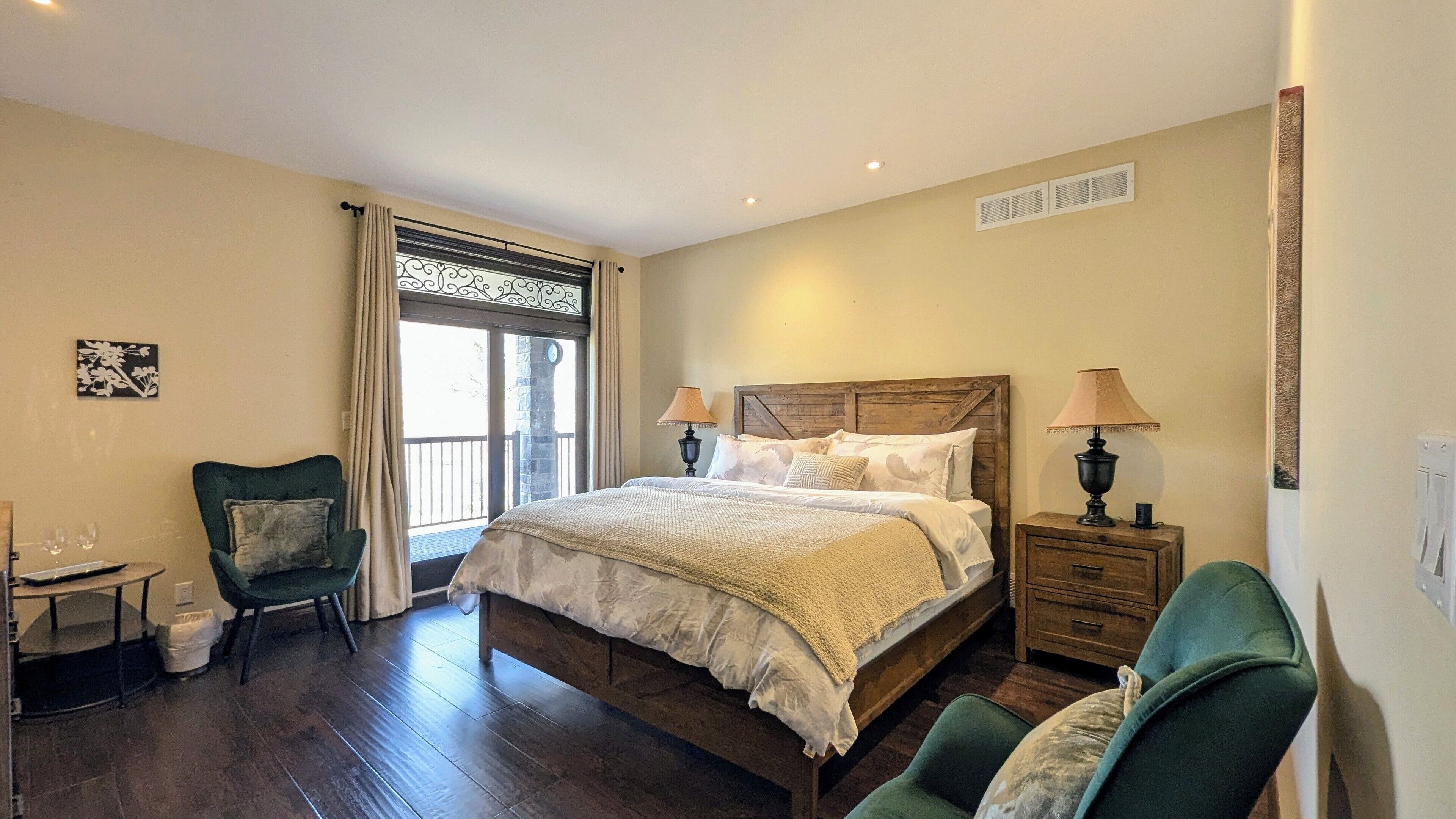 A bedroom with a large bed, two bedside tables with lamps, a chair, a small table with glasses, and a window with curtains.