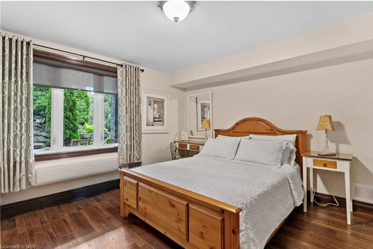 A bedroom with a wooden bed, a nightstand with a lamp, a window with curtains, and a mirror on the wall.