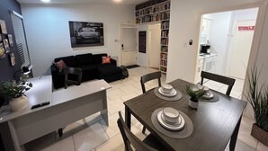 Een moderne woonkamer met een zwarte bank, een eettafel met borden en een plant, een plafondventilator en een boekenplank vol boeken.