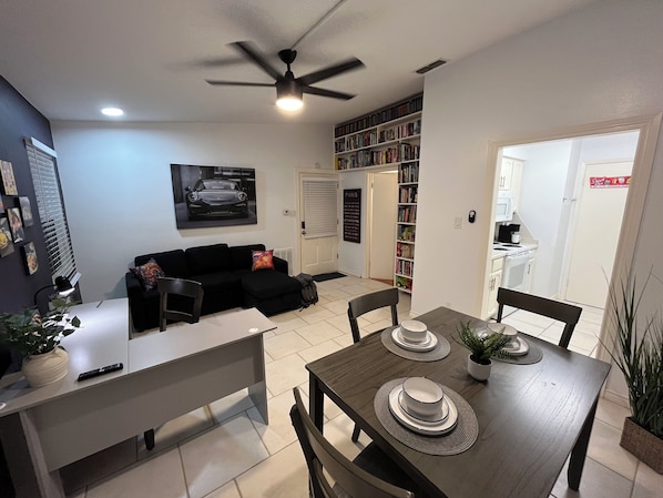 A modern living room with a black sofa, a dining table set with plates and a plant, a ceiling fan, and a bookshelf filled with books.