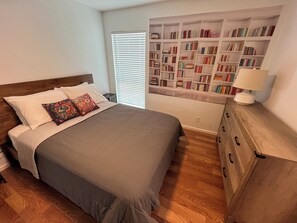 Een slaapkamer met een bed, nachtkastje, houten dressoir en een boekenplank vol boeken.