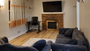 A living room with a fireplace, a flat-screen TV mounted above it, a sofa, a chair, and a window with curtains. - Convenient/family friendly home with yard driveway

 (Silver Spring)
