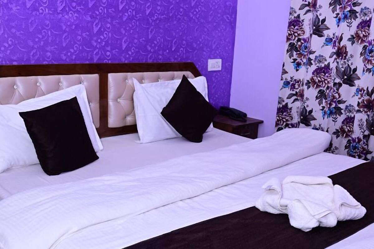 A hotel room with a large bed, two black and white pillows, a wooden headboard, a nightstand with a phone, and a floral curtain.