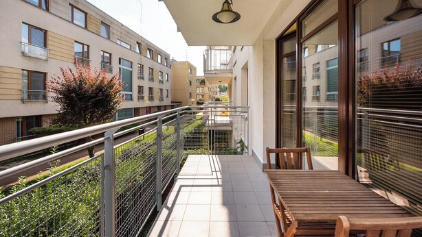 Een balkon met een houten tafel en stoelen, een metalen hekwerk en uitzicht op omliggende gebouwen.