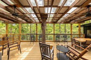 A covered outdoor area with wooden flooring, tables, chairs, and a central tree trunk. - Milam Ridge: Modern Cabin Escape. Hot Tub, Covered Deck, Fire Pit (Springfield)