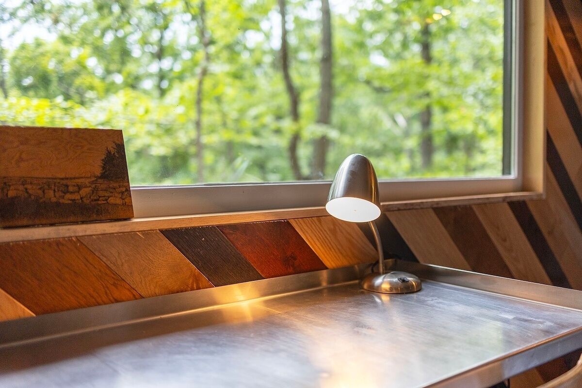 Un banco de madera con una mesa de metal y una lámpara, ubicado cerca de una ventana que ofrece vistas a árboles.