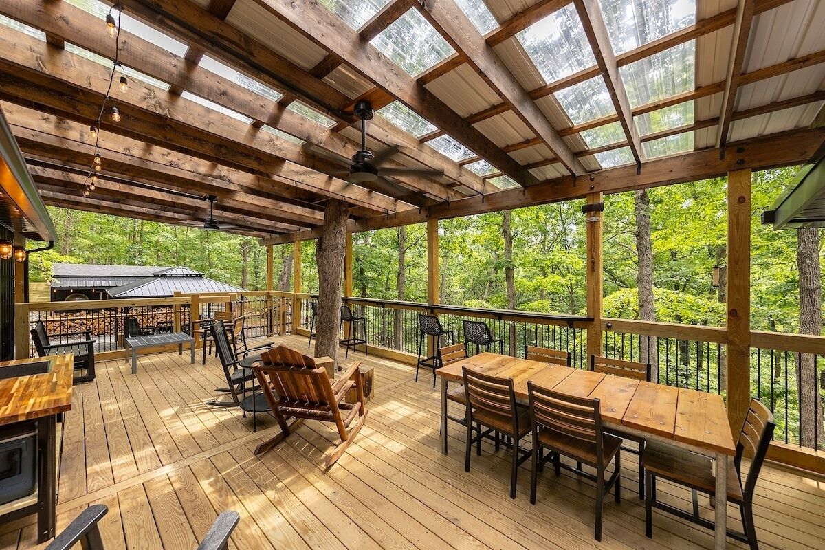 A wooden deck with tables and chairs, surrounded by glass panels and trees.