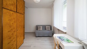 Une chambre avec une armoire en bois, un canapé gris, un bureau blanc et une fenêtre avec des rideaux blancs.