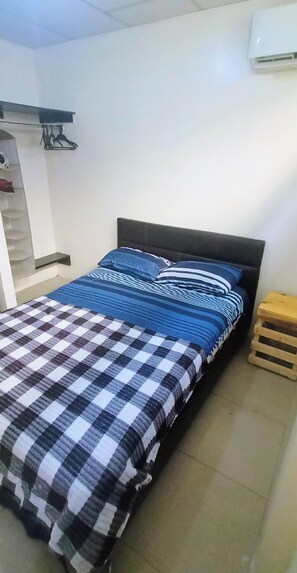 A bedroom with a bed, a wooden stool, a shelf with hanging items, and a wall-mounted air conditioner.