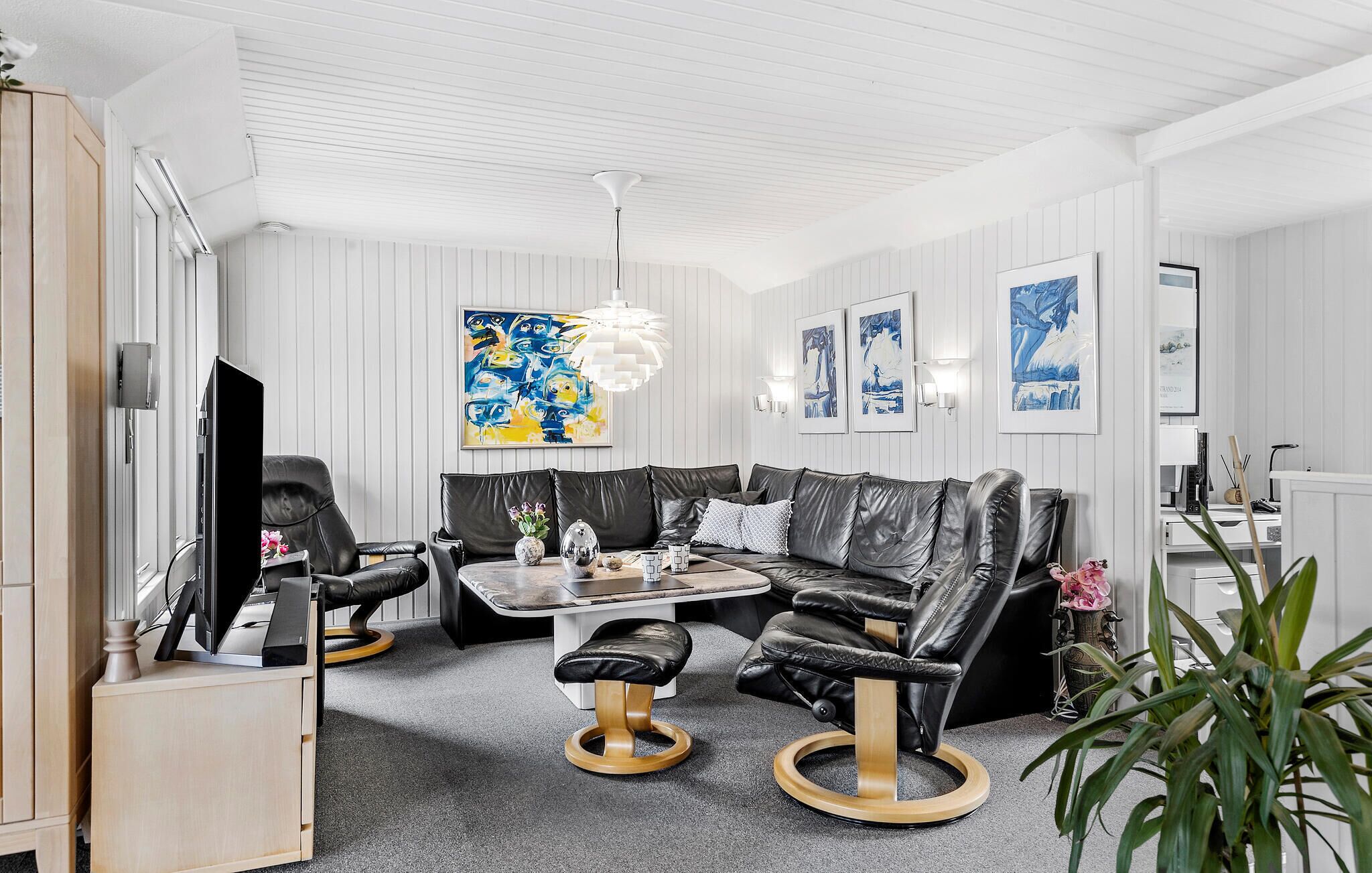 Een moderne woonkamer met een zwarte leren bank, een salontafel en een televisie. Er hangen kunstwerken aan de muur en in de hoek staat een plant.
