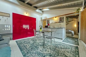 Une cuisine moderne avec des meubles rouges, un coin repas doté d’une table et de chaises en verre, et un escalier apparent menant à une chambre.