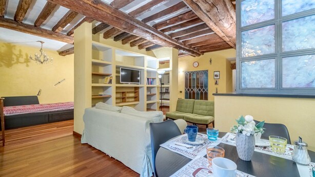 A cozy room with a wooden ceiling, a sofa, a green armchair, a dining table with newspapers, and a TV on a shelf.