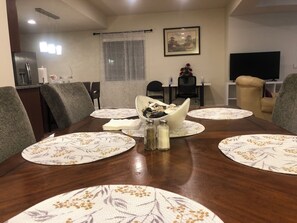 A dining room with a wooden table set for six, surrounded by chairs. There is a window with sheer curtains, a framed picture on the wall, and a television in the background. - Charming 2-bedroom basement Apartment with AC and WiFi in terrific Aurora (Aurora)