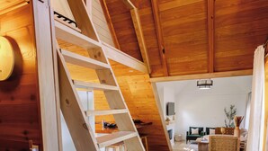 Een houten trap leidt naar een slaapkamer op een entresol, met uitzicht op de woonkamer beneden.