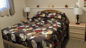 A bedroom with a bed, two bedside tables with lamps, a wall decoration, and a small shelf with a plant and a clock. - Sea Shell Inn. A cozy hideaway (Duncan)