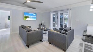 Un salon moderne avec deux canapés gris, un ventilateur de plafond, un téléviseur à écran plat et une vue dégagée sur l’extérieur grâce à de grandes portes vitrées.