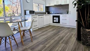 A modern kitchen with white cabinets, a microwave, an oven, and a dining area with a table and chairs. - Quantrill Avenure Retreat (Benalla)