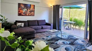 A modern living room with a grey sofa, a coffee table, and a large painting on the wall. There is a sliding glass door leading to an outdoor area with a table and chairs. - Quantrill Avenure Retreat (Benalla)