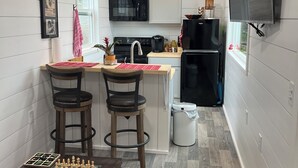 A compact kitchen with white paneling, a microwave, a refrigerator, and a sink. There are two bar stools and a small table with a chessboard.