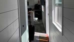 A narrow hallway with white walls and a wooden floor leading to a kitchen area with modern appliances and a window.