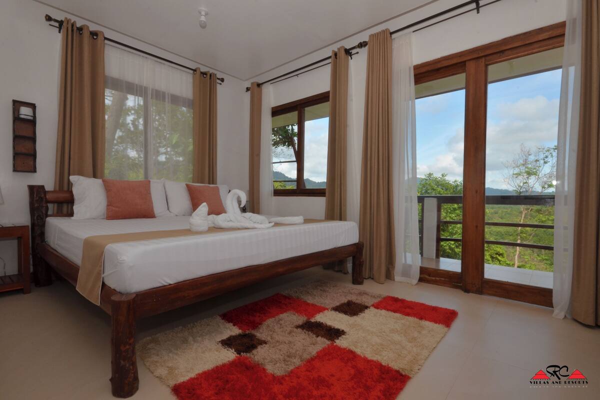 A bedroom with a large bed, a wooden headboard, and a view of greenery through large windows.