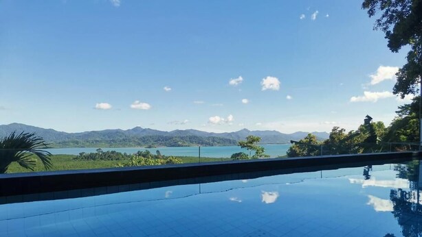 On peut clairement voir une piscine dont le fond est recouvert de motifs de carreaux, entourée d’une végétation luxuriante et avec une chaîne de montagnes qui se dessine à l’horizon.