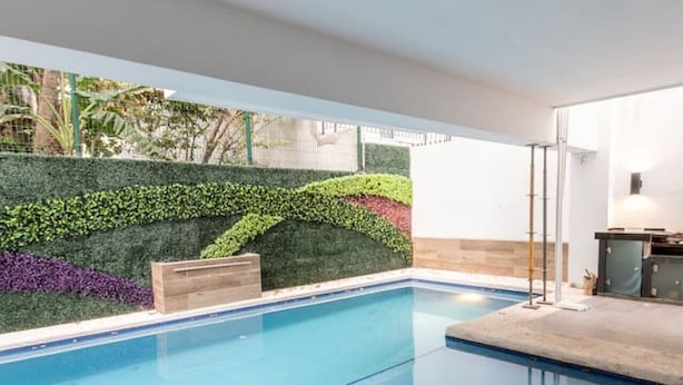 Una piscina con superficie de agua azul cristalina, rodeada por un jardín ajardinado con diversas plantas y una terraza de madera.