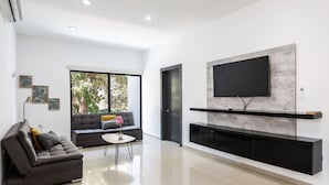 Una sala de estar moderna con un televisor de pantalla plana montado en la pared, un sofá gris, una pequeña mesa de centro con un jarrón y un ventanal con vistas a la vegetación.