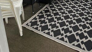 A covered outdoor seating area with a black and white checkered rug, a white plastic chair, and a wicker sofa with cushions.