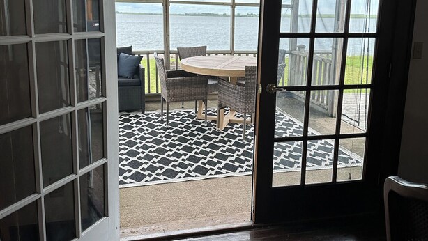A room with a large sliding glass door leading to a patio with a round table and chairs, a view of the water, and a patterned rug on the floor.