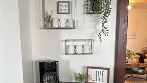 A dark wooden cabinet with a coffee maker on top, a framed sign saying "LIVE", and two shelves with white mugs and a small plant.