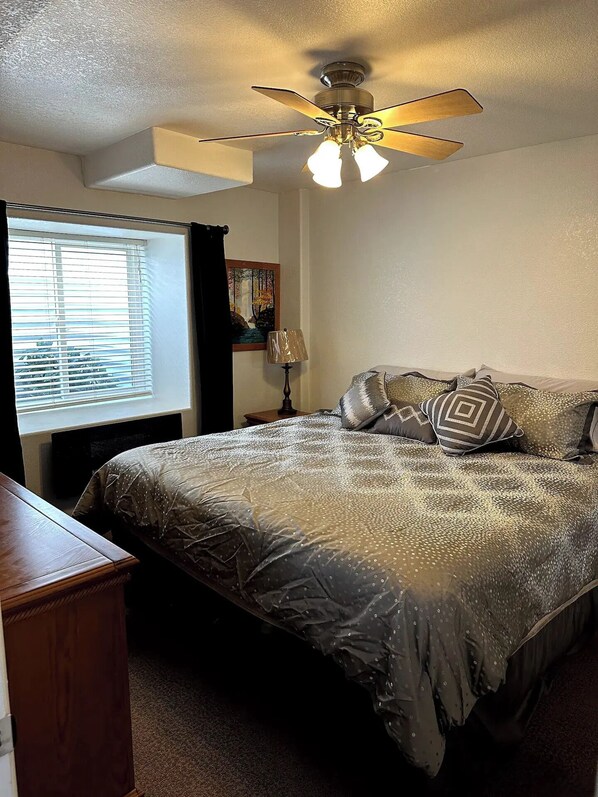A bedroom with a bed, a ceiling fan, a window with blinds, a lamp, and a painting on the wall.