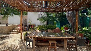 Une structure en bois formant une pergola, avec une table et des chaises en dessous, entourée de plantes en pot et d’un mur de pierre.