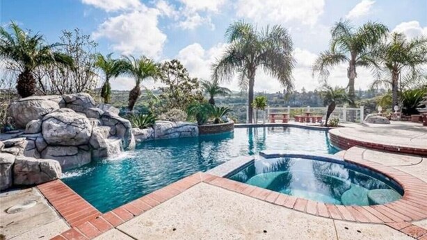 A swimming pool with a waterfall feature, surrounded by rocks and palm trees, with a view of the landscape in the background. - Luxury Hill Villa with Spectacular View (Anaheim)