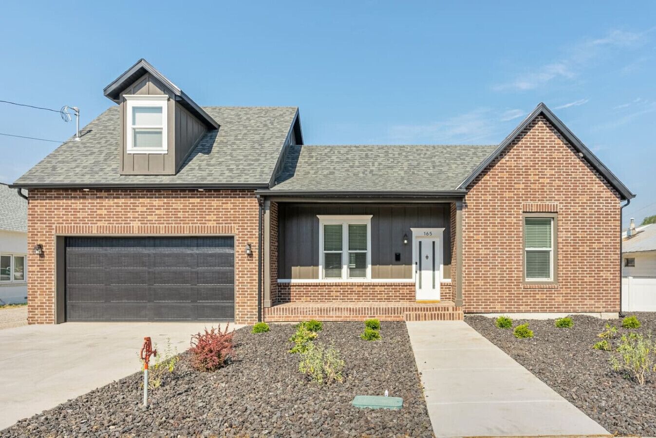 A two-story brick house with a dark gray roof, a garage, and a front porch with a small garden.