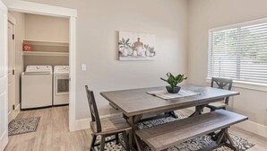 A dining area with a table, chairs, a bench, a potted plant, a painting, and a laundry room with appliances. - New! Main Street Retreat | BBQ | Pac-Man (Lehi)