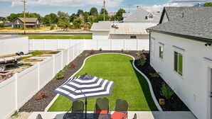 A backyard with a lawn, a patio with chairs and an umbrella, and a white fence. - New! Main Street Retreat | BBQ | Pac-Man (Lehi)