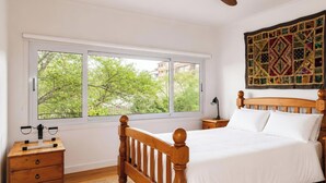 A bedroom with a wooden bed, a nightstand, a desk with a chair, a window with a view of trees, and a ceiling fan. - Caves Cottage - gorgeous 180 degree ocean views (Caves Beach)