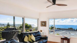 A living room with a black leather sofa, a ceiling fan, and large windows offering a view of the ocean and solar panels. - Caves Cottage - gorgeous 180 degree ocean views (Caves Beach)