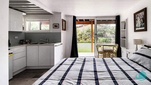 A bedroom with a bed, a small kitchenette, a sliding glass door to a balcony, and a view of greenery outside.