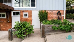 A residential property with a brick exterior, a white door, and a small window. There are wooden raised beds with green plants and a hose on the right side.