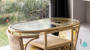 A corner table with a glass top and wicker frame, positioned near a window with a view of greenery.