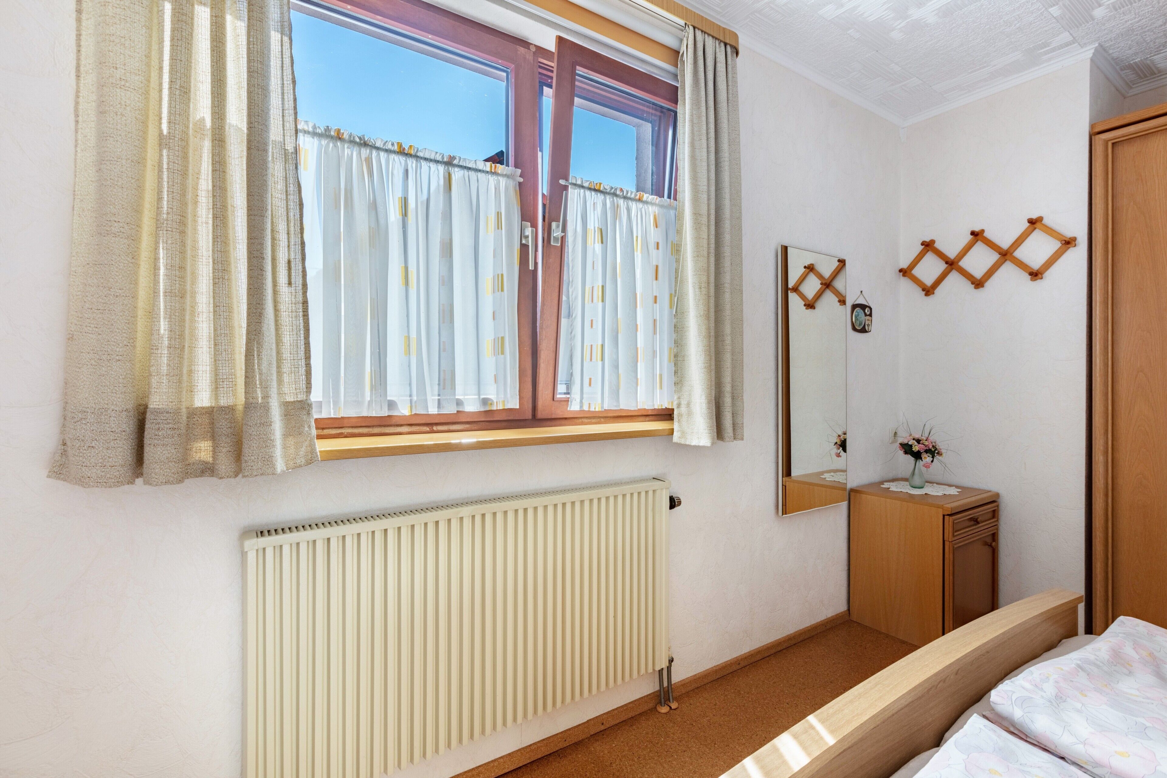Een slaapkamer met raam, radiator, nachtkastje van hout, spiegel en een bed.