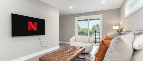 A modern living room with a flat-screen TV mounted on the wall, a brown leather bench, a white sofa with a fur throw, and a view of the outdoors through a sliding glass door.