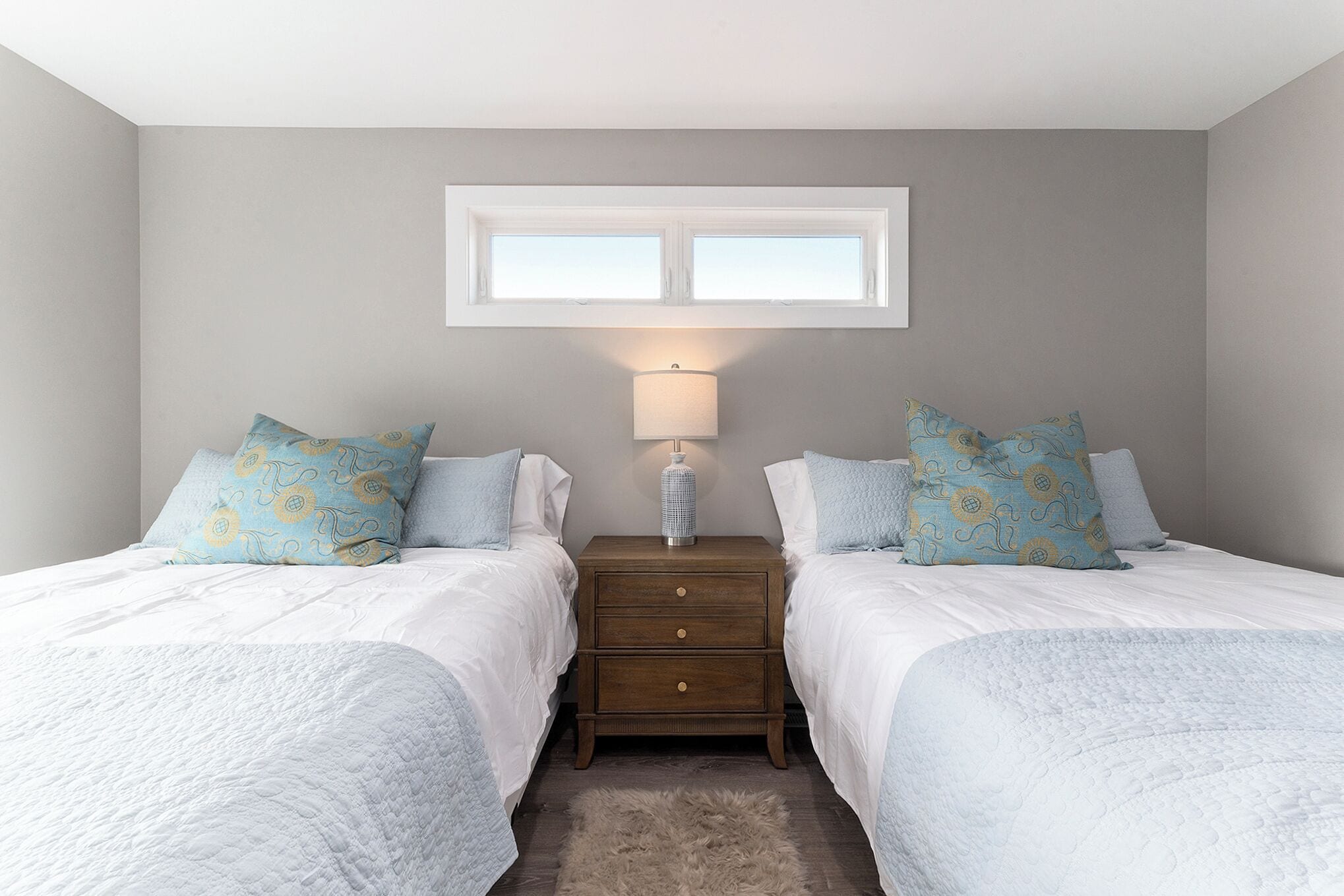A bedroom with two beds, a nightstand with a lamp, a window above the beds, and a fluffy rug on the floor.