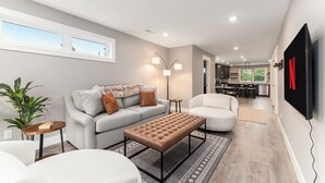 Ein modernes Wohnzimmer mit Sofa, Sessel und Hocker, einem Essbereich und einer Küche im Hintergrund.
