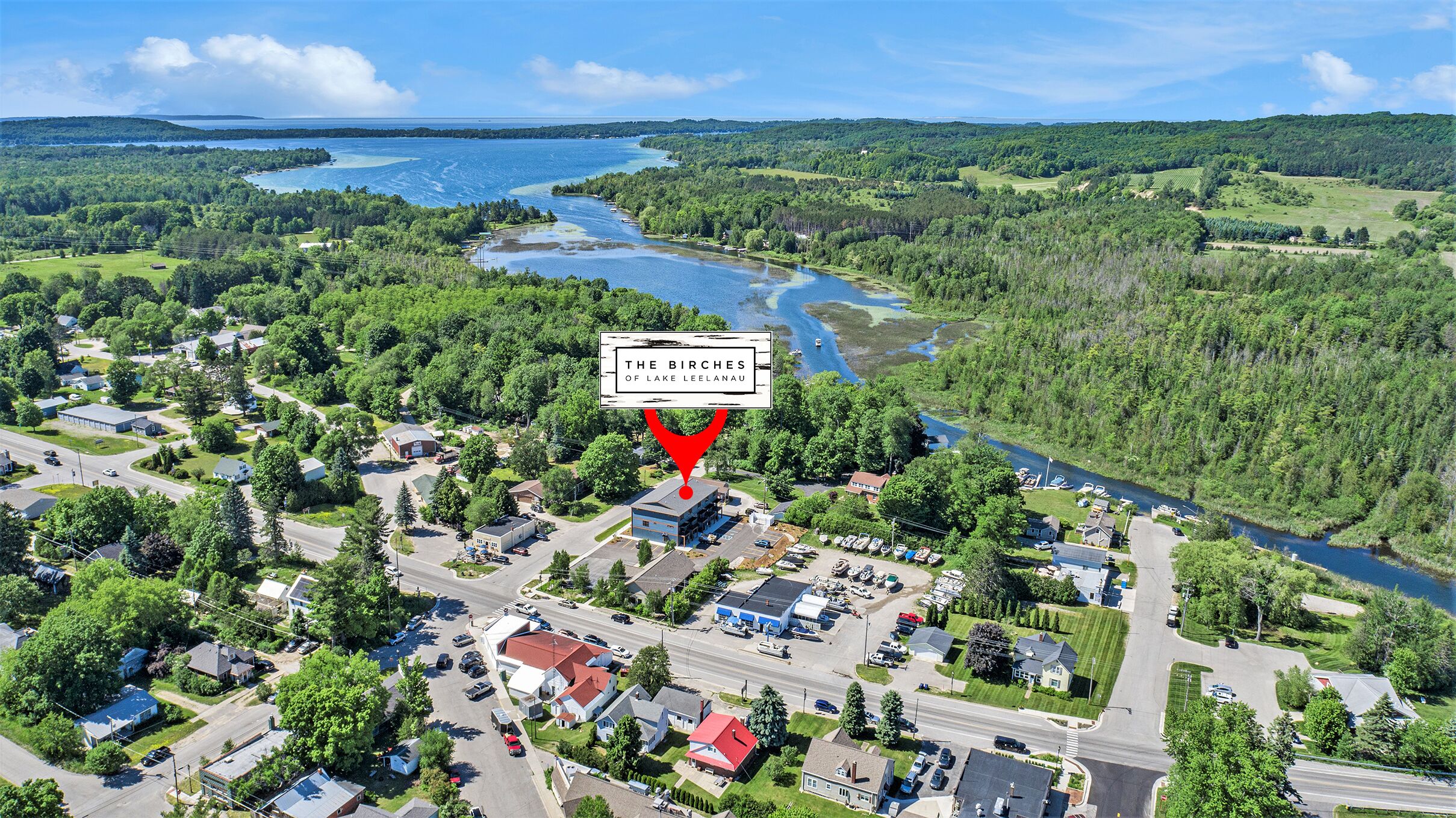 An aerial view of a small town with a prominent building labeled "The Birches of Lake Leelanau," surrounded by lush greenery and a lake.